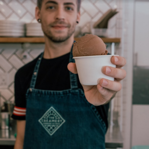 A guy in a Creamery apron holding a scoop of ice-cream toward the camera