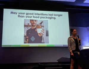 Cat speaking into mike in front of large slide which says: May your good intentions last longer than your packaging