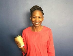 Photo of a smiling Phindi Dlamini, standing in front of a blue wall holding a Kraft Hot Cup